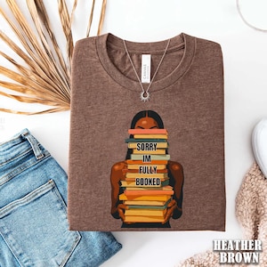 以下が含まれることがあります： 丸いネックラインのヘザーブラウンのTシャツ。シャツには、本を積み重ねて持っている人のグラフィックと「SORRY IM FULLY BOOKED」というテキストが描かれています。太陽のペンダントが付いたシルバーのネックレスも表示されています。