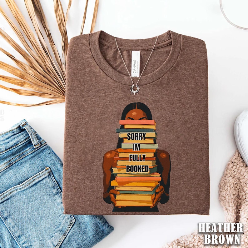 以下が含まれることがあります： 丸いネックラインのヘザーブラウンのTシャツ。シャツには、本を積み重ねて持っている人のグラフィックと「SORRY IM FULLY BOOKED」というテキストが描かれています。太陽のペンダントが付いたシルバーのネックレスも表示されています。