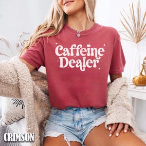 May include: Crimson-colored t-shirt with the text "Caffeine Dealer" in white, featuring a coffee cup graphic. The shirt is paired with denim shorts. A cream-colored jacket is draped over the arms.