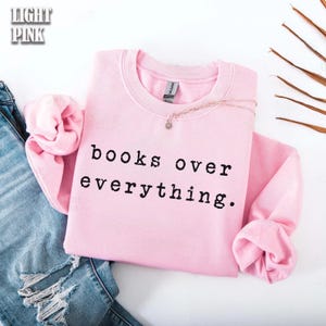 May include: Light pink sweatshirt with the text "books over everything" in black. A gold necklace with a small pendant rests on the sweatshirt. The sweatshirt is folded, with the sleeves tucked in. The words "light pink" are in the upper left corner.