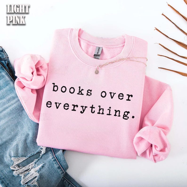 May include: Light pink sweatshirt with the text "books over everything" in black. A gold necklace with a small pendant rests on the sweatshirt. The sweatshirt is folded, with the sleeves tucked in. The words "light pink" are in the upper left corner.