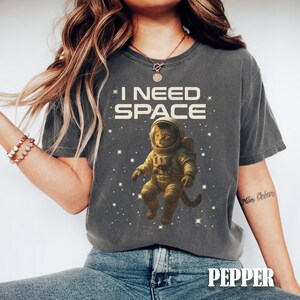 May include: Dark gray t-shirt with the text "I NEED SPACE" and a graphic of a cat in an astronaut suit floating in space. The word "PEPPER" is printed in white at the bottom of the shirt.