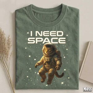May include: Moss green t-shirt with the text "I NEED SPACE" and a cartoon cat astronaut floating in space. The cat is wearing a spacesuit and helmet, surrounded by stars.