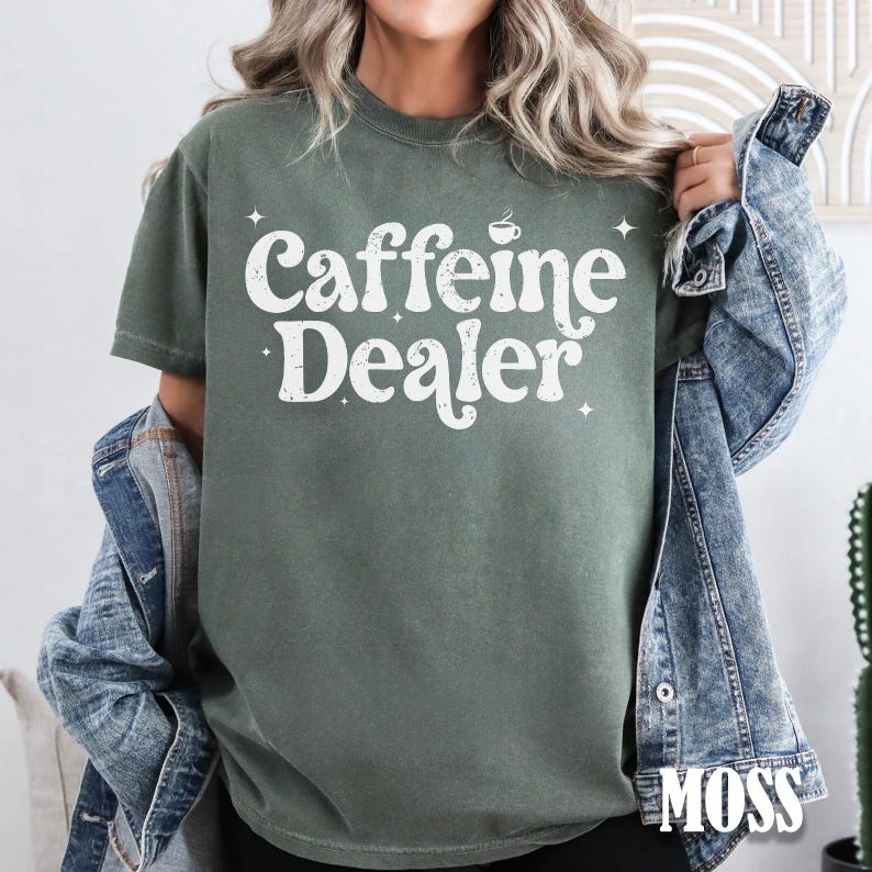 May include: A moss green t-shirt with the text "Caffeine Dealer" in white, featuring a coffee cup graphic. The shirt is a short-sleeved crew neck style.