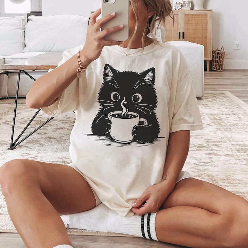 Peut inclure: T-shirt cr&egrave;me avec un chat noir tenant une tasse de caf&eacute;. Le chat a de grands yeux ronds et une expression mignonne. Le t-shirt est ample, parfait pour un style d&eacute;contract&eacute;.