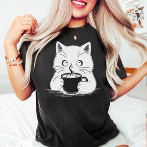 Peut inclure: T-shirt noir avec un dessin de chat blanc tenant une tasse de caf&eacute; noire. Le chat a de grands yeux et des moustaches, avec de la vapeur qui s'&eacute;chappe du caf&eacute;. Le t-shirt est de style d&eacute;contract&eacute;, adapt&eacute; au quotidien.