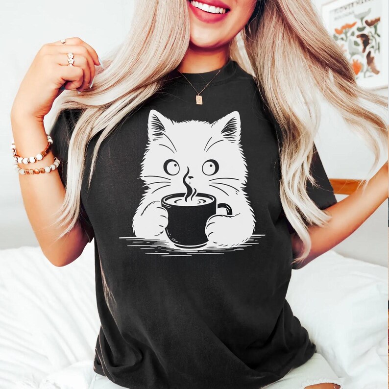 Peut inclure: T-shirt noir avec un dessin de chat blanc tenant une tasse de caf&eacute; noire. Le chat a de grands yeux et des moustaches, avec de la vapeur qui s'&eacute;chappe du caf&eacute;. Le t-shirt est de style d&eacute;contract&eacute;, adapt&eacute; au quotidien.