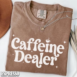 May include: Espresso-colored t-shirt with the words "Caffeine Dealer" in white, along with a coffee cup graphic. The shirt has a crew neck and short sleeves. A silver necklace with a small pendant is draped across the shirt.