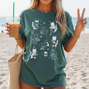 May include: Dark teal t-shirt featuring a white illustration of various cats in different poses, some holding coffee cups and milk cartons. The cats are depicted with star accents. The shirt is made of a soft material.