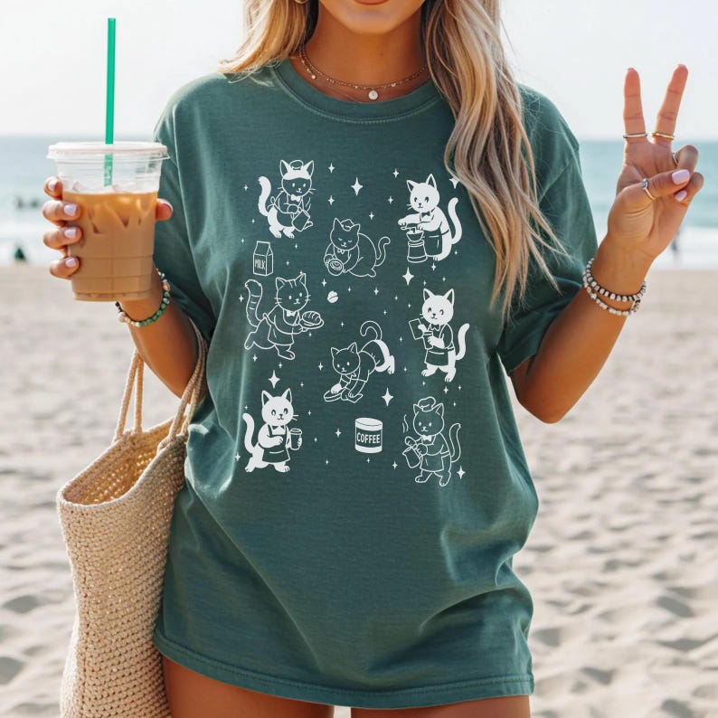 May include: Dark teal t-shirt featuring a white illustration of various cats in different poses, some holding coffee cups and milk cartons. The cats are depicted with star accents. The shirt is made of a soft material.