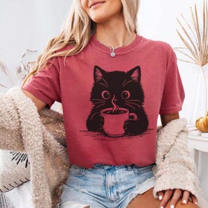 Peut inclure: T-shirt bordeaux avec un dessin de chat noir buvant du caf&eacute;. Le chat a de grands yeux et des moustaches. Le t-shirt est &agrave; manches courtes et de coupe d&eacute;contract&eacute;e. Le style est d&eacute;contract&eacute; et confortable.