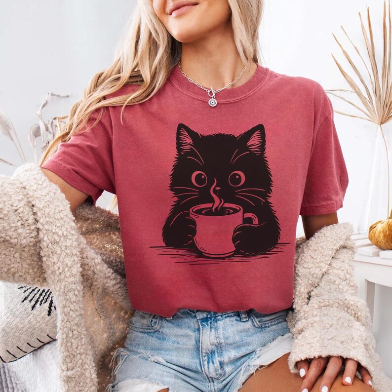 Peut inclure: T-shirt bordeaux avec un dessin de chat noir buvant du caf&eacute;. Le chat a de grands yeux et des moustaches. Le t-shirt est &agrave; manches courtes et de coupe d&eacute;contract&eacute;e. Le style est d&eacute;contract&eacute; et confortable.
