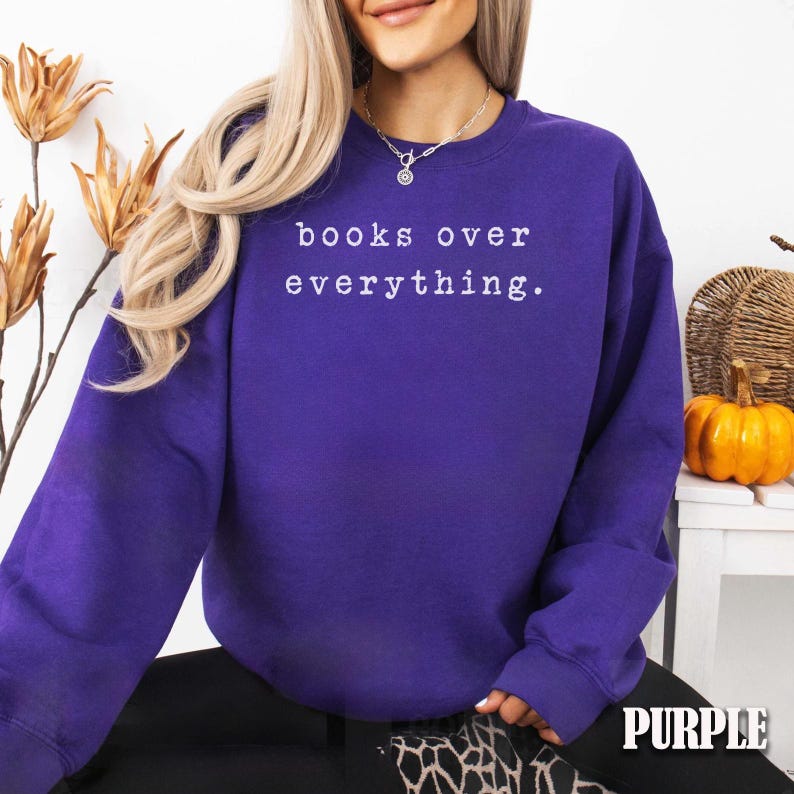 May include: A purple sweatshirt with the text "books over everything" in white. The sweatshirt is a vibrant shade of purple and is made of a soft material. The image also includes a pumpkin and decorative flowers.