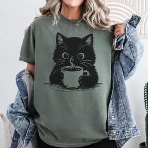 Peut inclure: T-shirt vert olive avec un motif de chat noir tenant une tasse de caf&eacute;. Le chat a de grands yeux et des moustaches, avec de la vapeur qui s'&eacute;chappe de la tasse. Le design est simple, de style dessin au trait, pour les amoureux des chats.