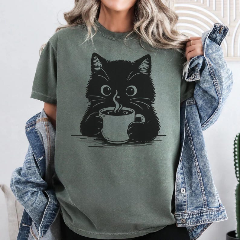 Peut inclure: T-shirt vert olive avec un motif de chat noir tenant une tasse de caf&eacute;. Le chat a de grands yeux et des moustaches, avec de la vapeur qui s'&eacute;chappe de la tasse. Le design est simple, de style dessin au trait, pour les amoureux des chats.