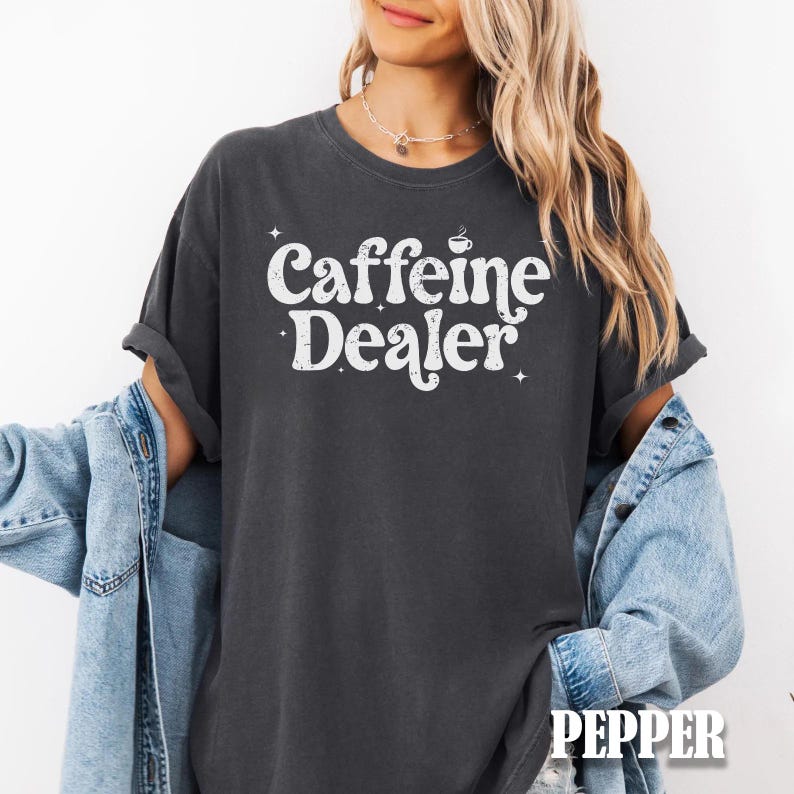May include: Dark gray t-shirt with the text "Caffeine Dealer" in white, featuring a coffee cup graphic. The shirt has rolled-up sleeves and a casual fit. The word "PEPPER" is printed at the bottom.