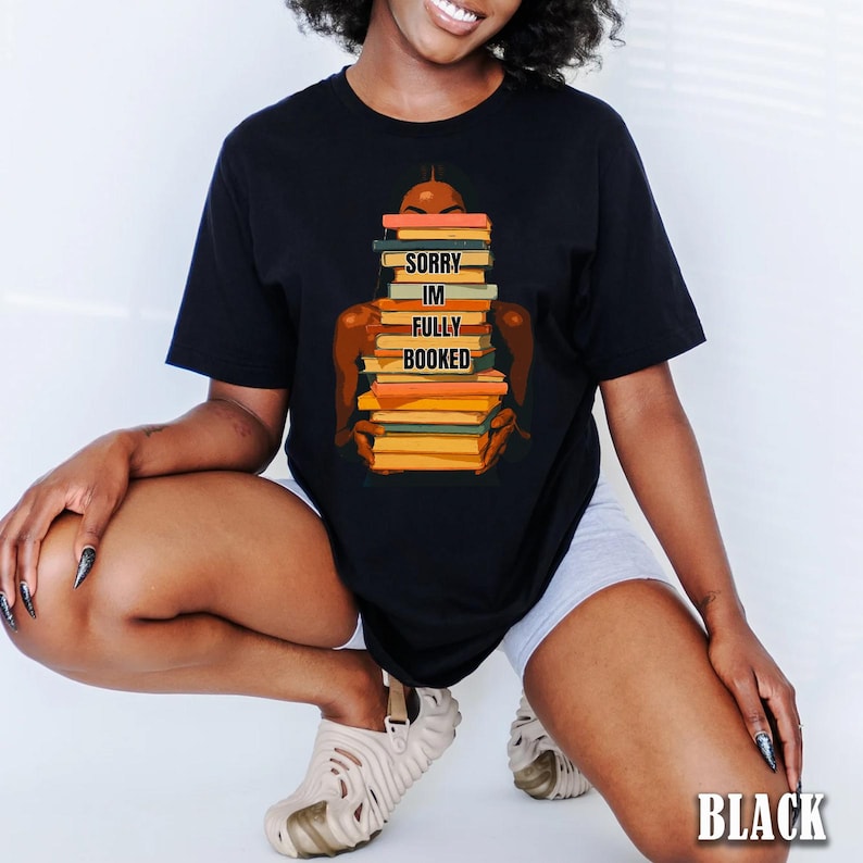 以下が含まれることがあります： 黒いTシャツ。本を抱える人のグラフィック。本はオレンジ、黄色、緑の色合い。本には「SORRY IM FULLY BOOKED」と書かれています。「BLACK」の文字が右下にあります。