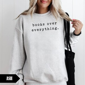 May include: Light gray sweatshirt with the text "books over everything" in black. The sweatshirt is a crew neck style and has long sleeves. A black tote bag is visible on the right side of the image.