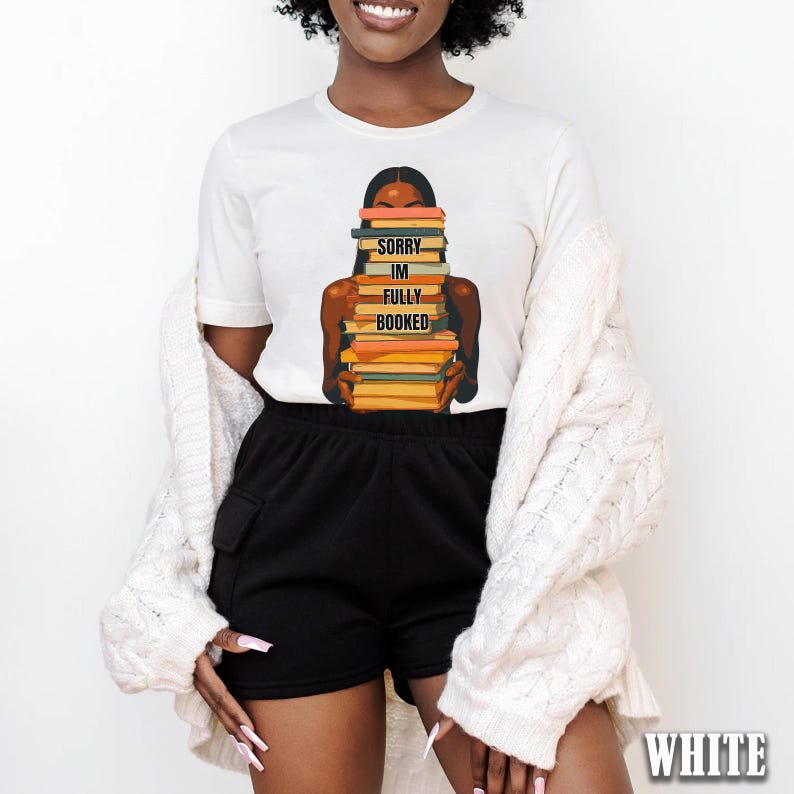 以下が含まれることがあります： 本を抱える人のグラフィックがプリントされた白いTシャツ。「SORRY IM FULLY BOOKED」の文字が本にプリントされています。その人は肌が黒く、黒髪です。このシャツは黒いショートパンツと白いカーディガンと組み合わせています。