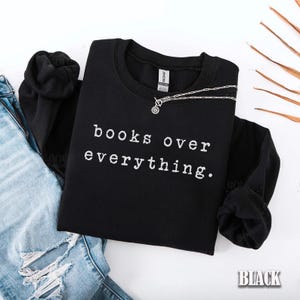 May include: Black sweatshirt with the text "books over everything" in white. A silver necklace with a circular pendant rests on the shirt. The word "BLACK" is in the bottom right corner. The sweatshirt is folded next to a pair of blue jeans.