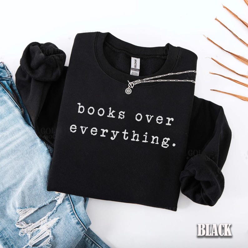 May include: Black sweatshirt with the text "books over everything" in white. A silver necklace with a circular pendant rests on the shirt. The word "BLACK" is in the bottom right corner. The sweatshirt is folded next to a pair of blue jeans.