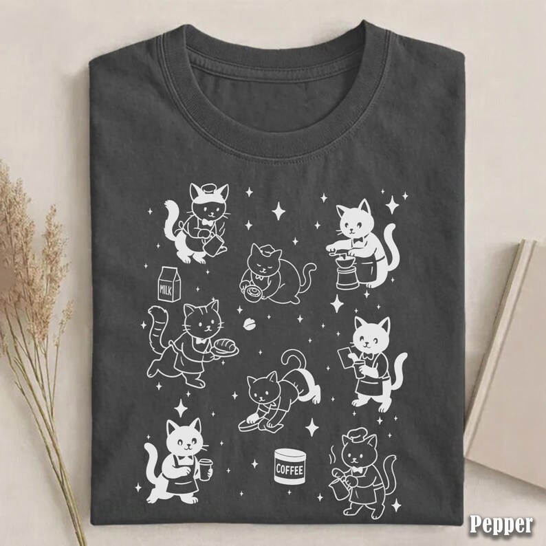 May include: Dark gray t-shirt featuring a white graphic of cartoon cats in various barista roles, such as pouring coffee and serving pastries. The word "Pepper" is printed in the bottom right corner.