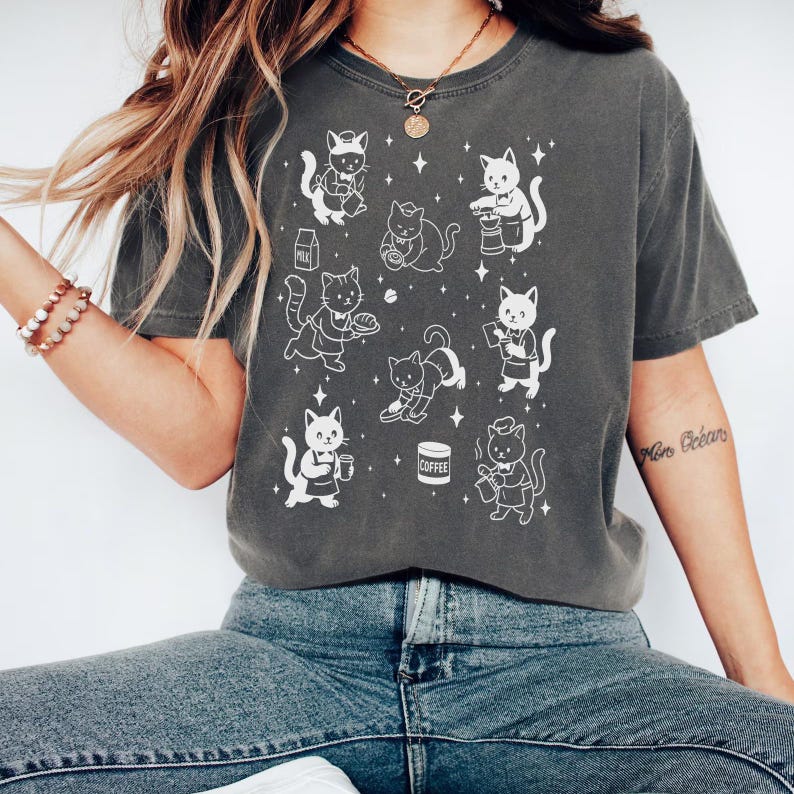 May include: Dark grey t-shirt featuring a repeating pattern of white cartoon cats in various poses, some holding food and drinks. The cats are depicted with chef hats and aprons. The shirt is a comfortable, casual style.