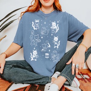 May include: A blue t-shirt featuring a white illustration of cats in various poses, some holding coffee cups and food items. The word "Coffee" is printed on a coffee cup. The shirt has short sleeves and a relaxed fit.
