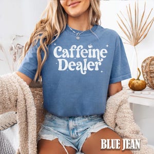 May include: A blue jean-colored t-shirt with the words "Caffeine Dealer" in white script. The shirt has a crew neck and short sleeves. The text is accompanied by a coffee cup graphic and star accents. The words "BLUE JEAN" are at the bottom of the image.