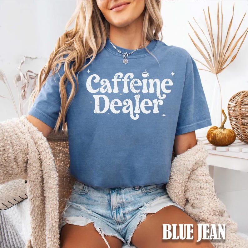 May include: A blue jean-colored t-shirt with the words "Caffeine Dealer" in white script. The shirt has a crew neck and short sleeves. The text is accompanied by a coffee cup graphic and star accents. The words "BLUE JEAN" are at the bottom of the image.