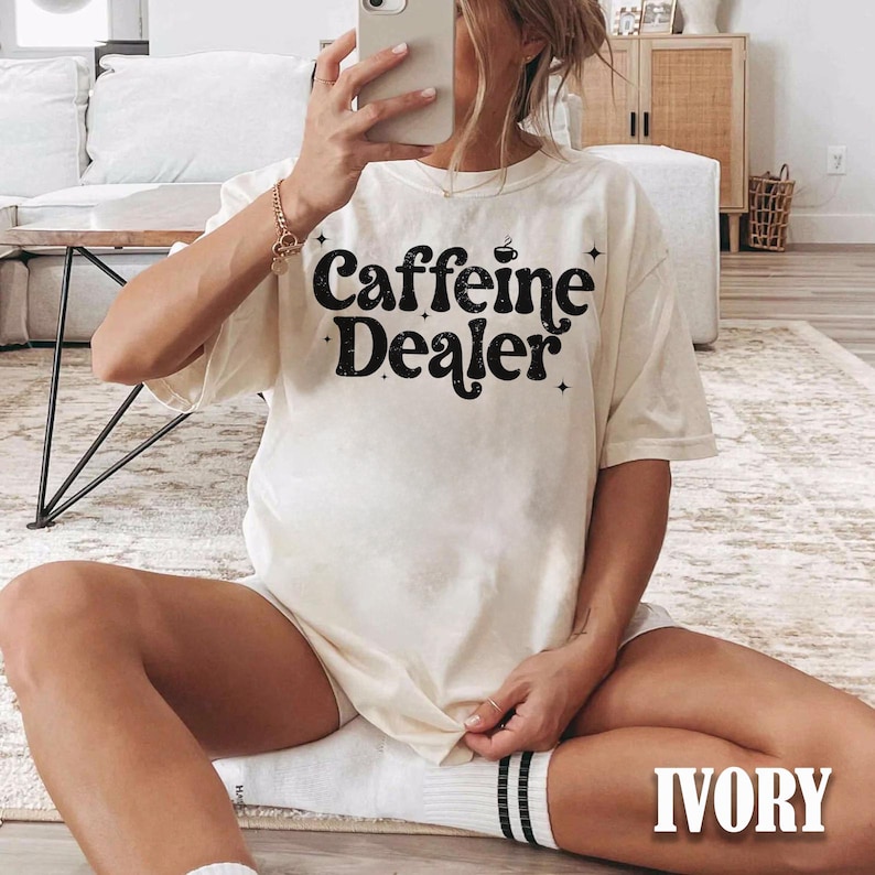 May include: Ivory-colored t-shirt with the text "Caffeine Dealer" in a black, retro font. The shirt has a relaxed fit and short sleeves. The image shows the shirt being worn.