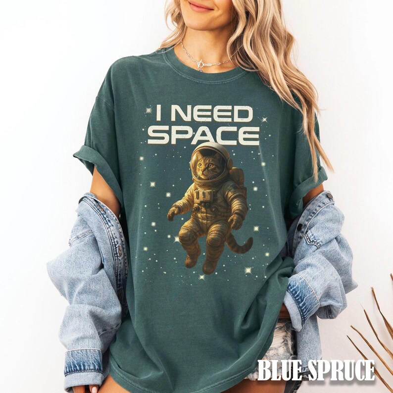 May include: A teal-colored t-shirt with the text "I NEED SPACE" in white block letters. The shirt features a graphic of a cat in an astronaut suit floating in space among stars. The words "BLUE SPRUCE" are at the bottom.