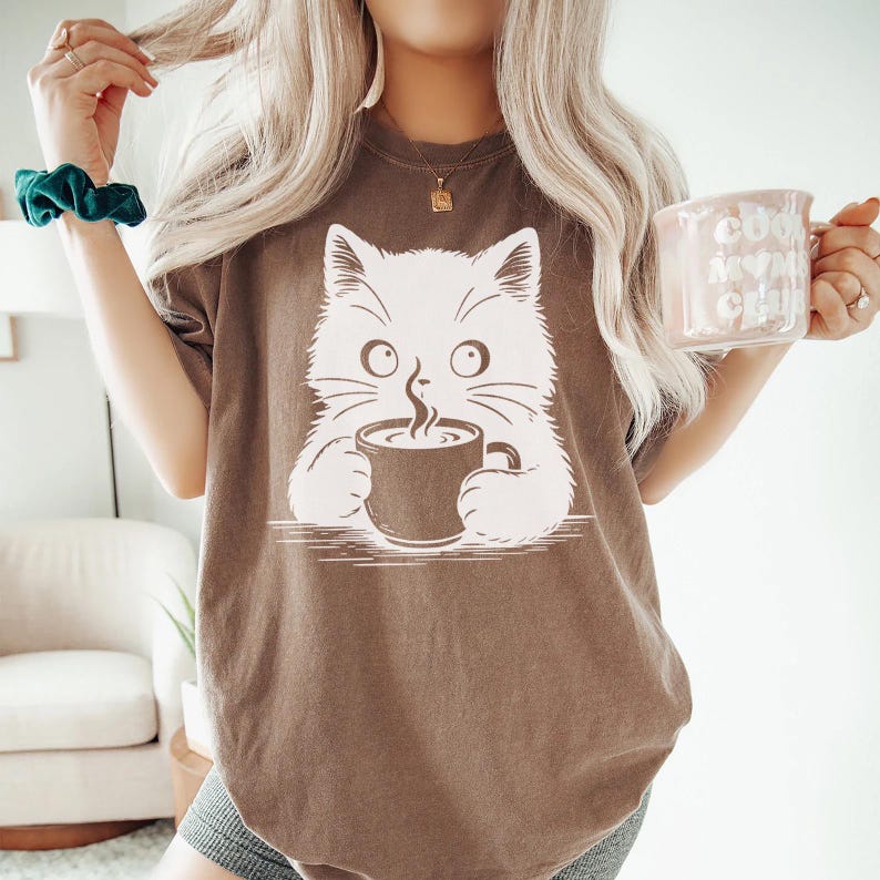 Peut inclure: T-shirt marron avec un motif de chat blanc tenant une tasse de caf&eacute;. Le chat a de grands yeux et de la vapeur s'&eacute;l&egrave;ve de la tasse. Une tasse rose avec le texte "Cool Moms Club" est tenue dans la main droite.