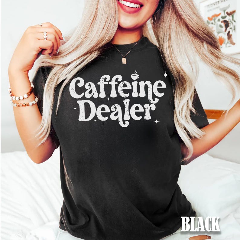 May include: Black t-shirt with the text "Caffeine Dealer" in white, stylized font. The design includes a small coffee cup icon above the word "Dealer" and star accents. The shirt is a solid black color.