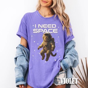 May include: Violet-colored t-shirt with the text "I NEED SPACE" above a cartoon cat astronaut floating in space. The cat is wearing a spacesuit and helmet, surrounded by stars.