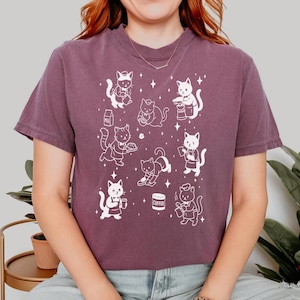 May include: A mauve t-shirt featuring a white graphic of cartoon cats in various poses, some holding coffee cups and baking. Other cats are shown with milk cartons and coffee beans. The shirt has a crew neck and short sleeves.