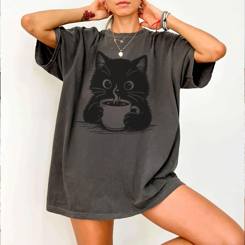 Peut inclure: T-shirt gris fonc&eacute; avec un motif de chat noir tenant une tasse de caf&eacute;. Le chat a de grands yeux et des moustaches. Le t-shirt a des manches courtes et une coupe d&eacute;contract&eacute;e, pour un usage quotidien.