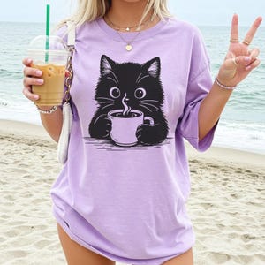 Peut inclure: T-shirt lilas avec un dessin de chat noir tenant une tasse de caf&eacute;. L'illustration du chat a de grands yeux et de la vapeur s'&eacute;chappe de la tasse. Le t-shirt est ample et &agrave; manches courtes.
