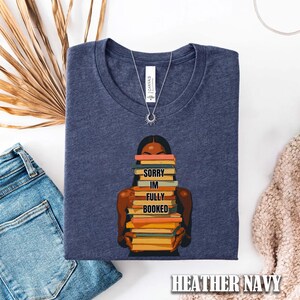 以下が含まれることがあります： 本を抱える人のグラフィックがプリントされた、ヘザーネイビーのTシャツ。「Sorry I'm Fully Booked」の文字が本に書かれています。太陽のペンダントが付いたシルバーのネックレスが見えます。Tシャツは、青いジーンズとベージュのセーターの隣に折りたたまれています。