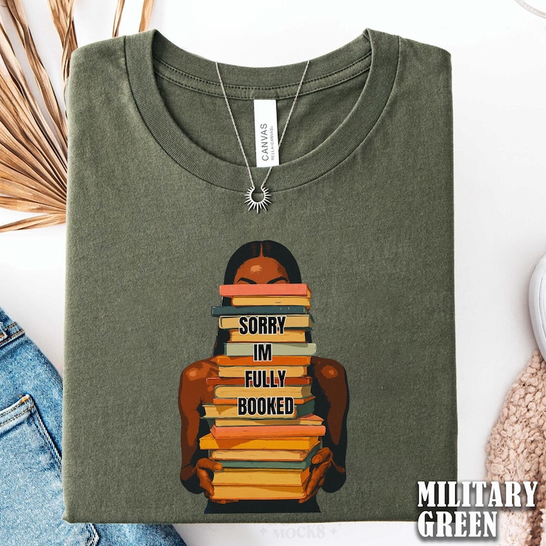 以下が含まれることがあります： ミリタリーグリーンのTシャツ。本を抱える人物のグラフィックが描かれています。本には「SORRY IM FULLY BOOKED」と書かれています。太陽のペンダントが付いたシルバーのネックレスが見えます。Tシャツは折りたたまれています。