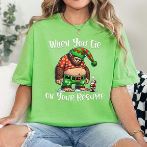 May include: Green t-shirt with a white graphic of a cartoon Yeti wearing a Santa hat and holding a gift. The text "When you lie on your resume" is printed on the shirt.