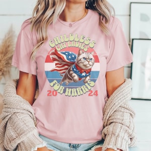 May include: Pink t-shirt with a graphic of a cat wearing a superhero cape and a red, white, and blue design. The text on the shirt reads "Childless Cat Ladies for Harris 2024".