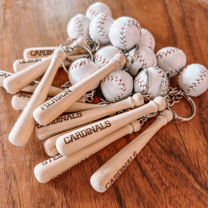 May include: A collection of baseball-themed keychains featuring miniature wooden bats and white leather baseballs with red stitching. Each wooden bat is engraved with the word Cardinals or a name such as Pierson or Olson, attached to a metal ring.