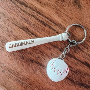 May include: A keychain featuring a white miniature baseball bat with the word CARDINALS engraved in brown, attached to a silver metal ring and chain with a small white baseball charm that has red stitching details throughout the surface.
