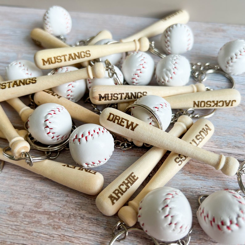 May include: A collection of baseball keychains featuring miniature wooden bats and white baseballs with red stitching. The bats are engraved with the text MUSTANGS, DREW, ARCHIE, and JACKSON. Each item includes a metal ring for attachment.