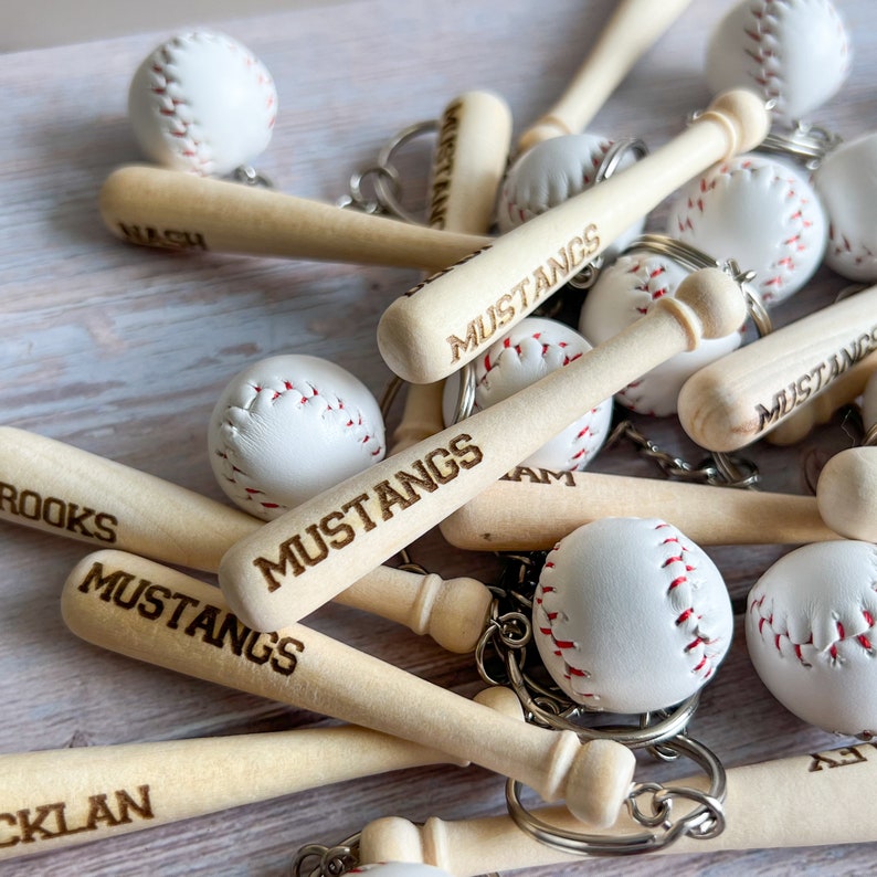 May include: A collection of keychains featuring miniature wooden baseball bats and white baseballs with red stitching. The bats are engraved with the text MUSTANGS, BROOKS, NASH, or CLAN. Each bat is attached to a metal keyring for attachment.