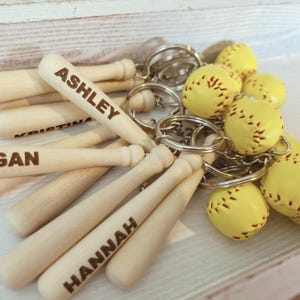 May include: A collection of keychains featuring miniature wooden baseball bats and yellow softballs with red stitching. Each bat is engraved with a name, including ASHLEY, KRISTINA, GAN, and HANNAH, attached to a silver metal ring.
