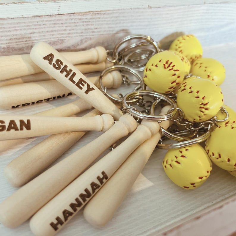 May include: A collection of keychains featuring miniature wooden baseball bats and yellow softballs with red stitching. Each bat is engraved with a name, including ASHLEY, KRISTINA, GAN, and HANNAH, attached to a silver metal ring.