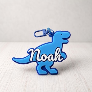 May include: A blue silicone keychain shaped like a dinosaur with the name Noah written in white cursive script across the center. The keychain features a matching blue lobster claw clasp attached to the top of the dinosaur figure.