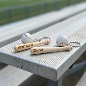May include: Two baseball-themed keychains, each featuring a miniature wooden baseball bat and a small white baseball with red stitching. One bat is engraved with the word MUSTANGS, and the other bat is engraved with the name DREW.